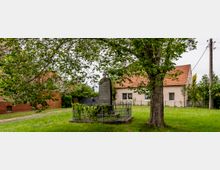 Denkmal auf einer grünen Wiese, umgeben von einem schmiedeeisernen Zaun, mit einem großen Baum im Vordergrund und Wohnhäusern mit roten Ziegeldächern im Hintergrund.