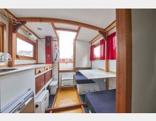 Innenraum eines kleinen Wohnwagens mit Holzelementen. Zu sehen sind eine Sitzgruppe mit blau gepolsterten Bänken und einem weißen Tisch, an den Seiten rote Vorhänge, ein kleiner Küchenbereich mit Spüle und Schrankfächern sowie eine schmale gelbe Bodenfläche.