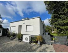 Flaches, modernes Einfamilienhaus mit weißer Fassade und Flachdach, umgeben von einem gepflasterten Hofbereich. Im Vordergrund befinden sich Pflanzen in Töpfen, eine Klimaanlage und ein grauer Zaun, rechts grenzt das Grundstück an eine grüne Gartenfläche mit Bäumen.