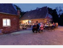 Ein beleuchteter Hof mit Kopfsteinpflaster und zwei Backsteingebäuden im traditionellen Stil. Mehrere Personen sitzen an Tischen mit Kerzen und Blumen, offensichtlich ein Abendessen oder eine Veranstaltung im Freien.