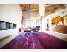 Ein modernes Wohnzimmer in einem Loft-Stil mit freiliegenden Ziegeldecken und sichtbaren Metallträgern. Der Raum enthält einen lila Teppich, einen schwarzen Sessel mit Hocker, eine Kaminwand, eine hölzerne Theke und große Fenster, die viel Licht hereinlassen.