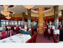 Elegantes asiatisches Restaurant mit kunstvoll gestalteten roten Holzmöbeln, großen Marmorsäulen und opulenten Kronleuchtern. Die Wände sind aufwendig dekoriert, mit traditionellen Mustern und Vasen, und ein großer, spiralförmiger Kristallleuchter dominiert die Mitte des Raumes.