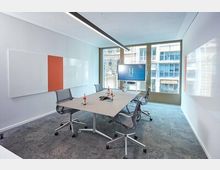 Moderner Meetingraum mit rechteckigem Tisch in der Mitte, umgeben von mehreren Bürostühlen. An der Wand befinden sich weiße und orangefarbene Tafeln sowie ein Bildschirm; ein großes Fenster bietet Blick auf städtische Gebäude.