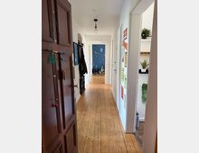 Langer, schmaler Wohnungsflur mit Holzdielenboden, weißen Wänden und einer einzelnen Deckenlampe. Links sind Garderobe und Spiegel neben einer dunklen Eingangstür, rechts ein Türrahmen mit Wandpostern und Regal mit Zimmerpflanzen; am Ende öffnet sich der Flur zu einem Raum mit dunkelblauer Wand und Fahrrad.