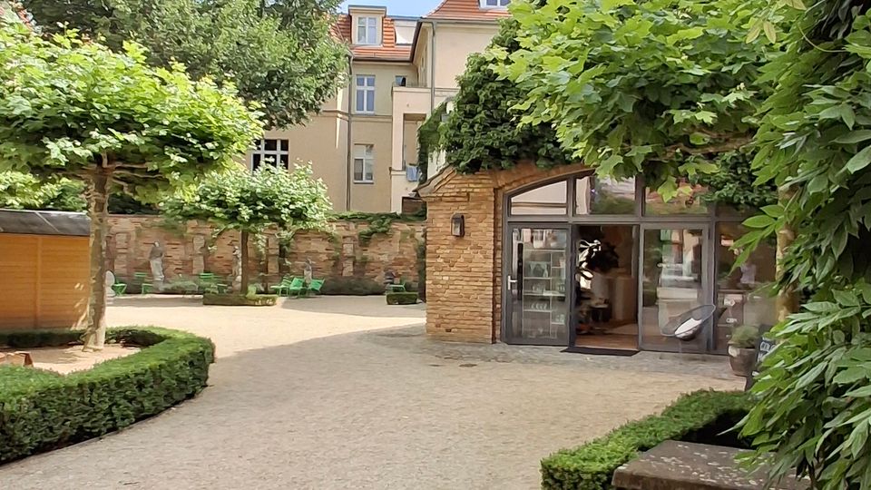 Innenhofgarten mit Kieswegen, niedrigen Buchshecken und dichtem Blätterdach aus Bäumen. Rechts steht ein kleiner Backsteinbau mit großer Glasfront, im Hintergrund eine Steinmauer und ein mehrstöckiges Wohnhaus.