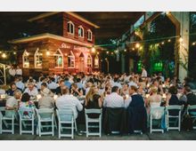 Eine große Gruppe von Menschen sitzt an langen, weiß gedeckten Tischen unter einer überdachten Außenfläche mit Lichterketten. Im Hintergrund steht ein rustikales, hölzernes Gebäude mit der Aufschrift „Oberhafenkantine“.