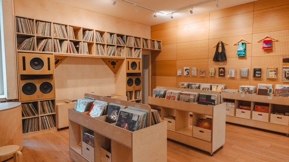 Interior of a modern record shop with light wood floors and walls, featuring built-in shelving filled with vinyl records and large speaker cabinets. Low display bins of records sit in the center, and a pegboard wall holds small merchandise and magazines under track lighting.