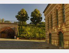 Historisches Gelände mit einem Backsteinbau auf der rechten Seite, dessen Fassade durch rote und gelbe Klinker sowie hohe Rundbogenfenster geprägt ist. Links ist eine massive Mauer mit Torbogen zu sehen, dahinter ein grasbewachsener Hang mit Bäumen.