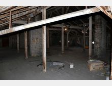 Ein leerer Dachboden mit Holzbalken und sichtbaren Stützpfeilern aus Holz und Beton. Der Boden ist aus Holzdielen und mit verstreuten Gegenständen, darunter eine Pappschachtel und Abfall, unordentlich belegt.