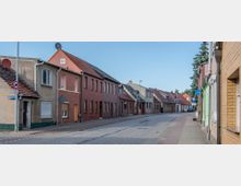 Eine ruhige Wohnstraße mit zweigeschossigen Häusern, die überwiegend aus Ziegel- und Putzfassaden bestehen, einige mit Satellitenschüsseln und Schrägdächern. Ein gepflasterter Gehweg und eine asphaltierte Fahrbahn verlaufen entlang der Häuser, die unter einem klaren blauen Himmel stehen.