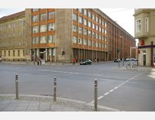 Straßenkreuzung in einer städtischen Umgebung mit einem mehrstöckigen Gebäude mit großer Fensterfront und gelblich-brauner Fassade im Hintergrund. Rechts im Bild befindet sich ein Eckhaus mit einer Apotheke und ein Fahrrad steht an einem Laternenpfahl.