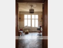 Ein heller Raum mit großen Sprossenfenstern und einer weißen Doppeltür, die zu einem Balkon führt. Der Raum hat einen Holzboden in Fischgrätmuster, eine abgenutzte Decke mit einem beschädigten Lampenschirm in der Mitte und enthält einfache Möbel, darunter ein Sofa, einen gelben Sitzpouf und einen Schreibtisch mit Stuhl.