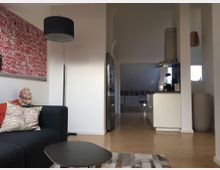 Wohnzimmer mit modernem Design, bestehend aus einem dunkelgrauen Sofa mit gemusterten Kissen, einem runden Couchtisch und einem Kunstwerk mit roten Schriftzügen an der Wand. Im Hintergrund befindet sich eine offene Küche mit weißer Einrichtung sowie ein Essbereich mit Holzboden und natürlichem Licht.