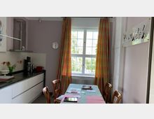 Helle Küche mit Esstisch im Vordergrund, bedeckt von einer Pastell-Tischdecke mit Arbeitsunterlagen. Hinten ein großes Fenster mit bunt karierten Vorhängen, daneben eine weiße Küchenzeile mit schwarzer Arbeitsplatte, Kaffeemaschine und Wanduhr.