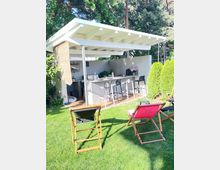 Moderne Gartenküche mit Überdachung, ausgestattet mit einer Theke, Barhockern und Küchenutensilien, umgeben von einer grünen Wiese und Bäumen im Hintergrund. Im Vordergrund stehen zwei Liegestühle auf dem Rasen.