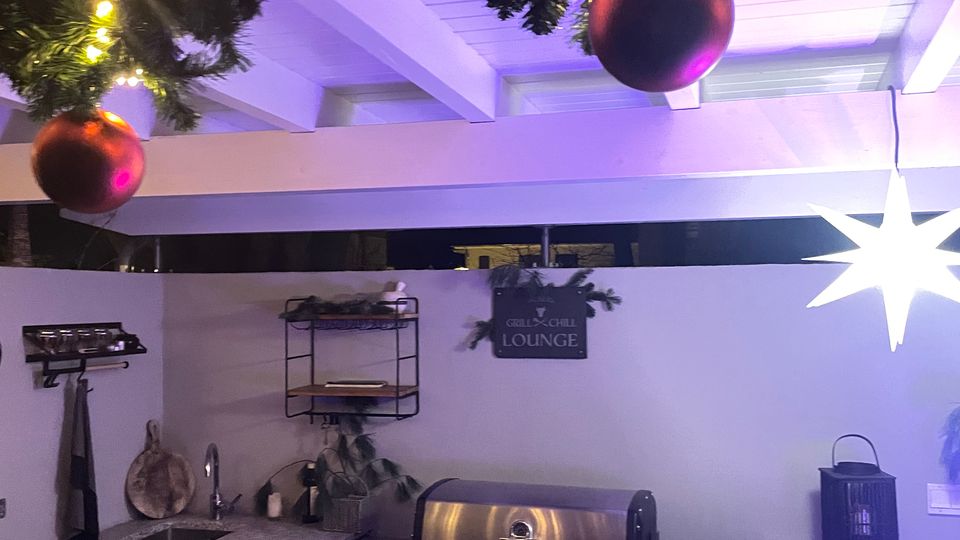 Outdoor-Küche mit einer Arbeitsplatte aus Granit, einem Edelstahlgrill, Regalen an der Wand und dekorativen Elementen. Weihnachtsdekoration mit Lichterketten, Tannenzweigen und Kugeln hängt von der Holzdecke.