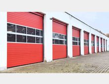 Außenansicht eines Gebäudes mit mehreren roten, rechteckigen Garagentoren, die mit waagerechten Linien unterteilt sind und teilweise verglaste Fensterreihen haben. Der Boden vor den Toren besteht aus grauen Pflastersteinen, und das Gebäude ist in einem schlichten Weiß gehalten.
