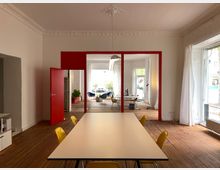 Moderner Innenraum eines Büros mit hohen Decken und Holzdielenboden. Im Vordergrund ein langer Konferenztisch mit gelben Stühlen, im Hintergrund rote Tür- und Fensterrahmen, die zu einem weiteren, hell erleuchteten Raum mit großen Fenstern und Sitzgelegenheiten führen.