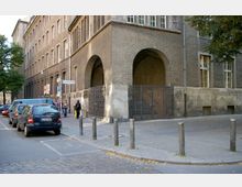 Straßenecke vor einem großen, historischen Gebäude mit grauer Fassade und Rundbogenöffnungen, die durch ein schmiedeeisernes Gitter verschlossen sind. Im Vordergrund sind geparkte Autos, ein Gehweg mit Passanten und Poller zu sehen; ein Straßenschild zeigt auf die Schönstedtstraße.