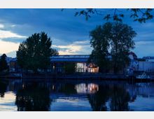 Gebäudekomplex am Wasser bei Dämmerung, mit großen Glasfassaden und beleuchteten Innenräumen, umgeben von Bäumen und einer Uferpromenade, die sich im ruhigen Wasser spiegelt. Im Hintergrund sind weitere Stadtgebäude sichtbar.