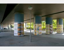 Ein Unterführungsbereich mit mehreren breiten, blauen Betonsäulen, die mit farbigen Plakaten beklebt sind. Pflastersteine bedecken den Boden, und im Hintergrund sind Bäume und parkende Autos zu sehen.