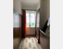 Schmaler Hausflur mit weißen Doppeltüren am Ende, die von einer orangefarbenen Vorhanghälfte und einem weißen Store bedeckt werden. Die Wände sind teils mit dunklem Holz vertäfelt, der Boden ist mit einem gemusterten Läufer ausgelegt, und auf der rechten Seite führt eine Treppe nach oben.