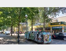 Straßenszene vor einem Bioladen in städtischer Umgebung. Im Vordergrund stehen mehrere Altglascontainer mit Plakaten, im Hintergrund gelbe Gebäude, Bäume und parkende Autos.