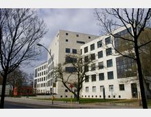 Modernes Bürogebäude mit heller Fassade und großen Fensterflächen, umgeben von Bäumen ohne Laub. Im Vordergrund befindet sich eine Grünfläche, im Hintergrund ein kleineres rotes Gebäude.