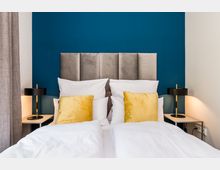 Modernes Schlafzimmer mit zwei nebeneinanderstehenden Betten, weißer Bettwäsche und goldfarbenen Zierkissen vor einem gepolsterten grauen Kopfteil. Dahinter eine dunkelblaue Akzentwand, links und rechts Nachttische mit schwarzen Tischlampen und ein grauer Vorhang am Fenster.