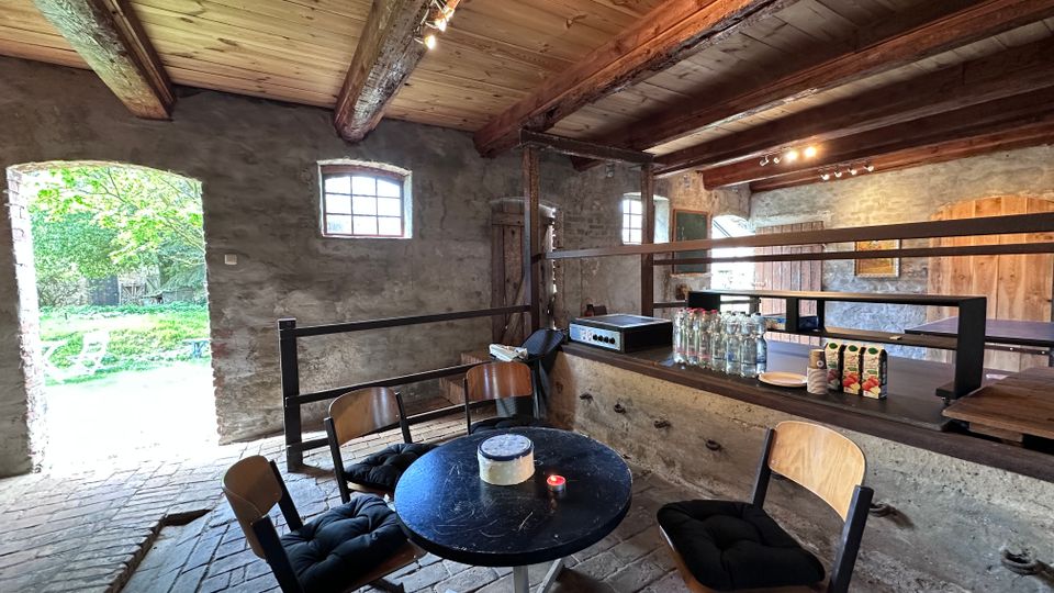 Rustikaler Innenraum eines umgebauten Scheunengebäudes mit groben Steinwänden, sichtbaren Holzbalken und Ziegelboden. Vor einem breiten Tresen mit Getränken stehen ein runder Tisch und mehrere Stühle; links öffnet sich ein großes Tor ins Grüne und kleine Fenster lassen Tageslicht herein.