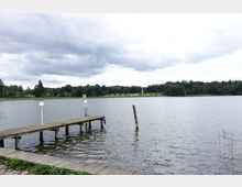 Holzsteg mit weißen Laternen über einem ruhigen See, im Hintergrund eine bewaldete Uferlandschaft und ein Obelisk auf einer freien Wiese unter einem bewölkten Himmel.