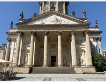 Fassade eines historischen Gebäudes mit klassizistischer Architektur, bestehend aus Säulenreihen, Reliefs und Skulpturen auf dem Giebel sowie in Nischen. Im Vordergrund ein gepflasterter Platz mit Tischen und Stühlen unter Sonnenschirmen.