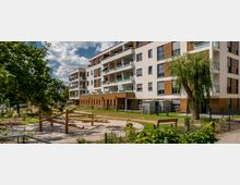 Mehrstöckiges modernes Wohngebäude mit weißen Wänden und Holzvertäfelung, umgeben von einer Grünfläche mit Spielplatz aus Holz, Wegen, einzelnen Bäumen und Bänken. Im Vordergrund ist ein umzäunter Bereich mit sandigem Boden und Klettergerüst zu sehen.