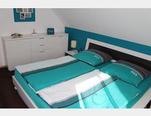 Schlafzimmer mit einem Doppelbett mit türkis-weißer Bettwäsche, einer weißen Kommode mit Schubladen und Deko-Elementen darauf, sowie einer Nachttischlampe auf einem kleinen Beistelltisch. Die Wand hinter dem Bett ist in Türkis gestrichen, während die restlichen Wände weiß sind.