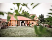 Fachwerkhof, umgeben von einer Wiese, mit einem großen Scheunengebäude mit rot gedecktem Dach und geöffneten Holztoren; im Vordergrund stehen Tische und Stühle, während im Hintergrund einige Personen stehen.