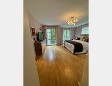 Ein geräumiges Schlafzimmer mit Holzboden, einem großen Doppelbett und einer modernen Hängeleuchte an der Decke. Die Rückwand hat zwei große Fenster mit Vorhängen, die Blick auf grüne Vegetation bieten, und eine Kommode mit einem Fernseher steht an der linken Wand.