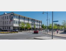 Kreisverkehr in einer Stadt mit einem modernen, weißen Geschäftsgebäude im Hintergrund und mehreren Geschäftsschildern, darunter „Woolworth“ und „TEDi“. Im Vordergrund sind eine bepflanzte Verkehrsinsel, parkende Autos, Fußgänger sowie Straßenlaternen und Bäume entlang des Gehwegs sichtbar.