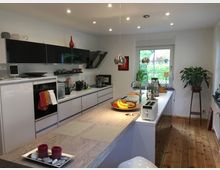 Moderne Küche mit heller Arbeitsfläche, darunter weißen Hochglanzschränken und schwarzen Hängeschränken. Ein großer Esstisch mit Holzoptik steht im Vordergrund, und ein Fenster mit Blick auf einen grünen Garten befindet sich im Hintergrund; zusätzlich sind Pflanzen und Küchenutensilien verteilt.