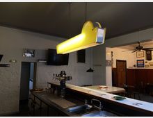 Ein rustikales Gasthaus mit Thekenbereich, Zapfhähnen und gelber Hängelampe. Weiße Steinwandoptik, dunkle Holzmöbel, Deckenventilator und verschiedene dekorative Elemente wie Bilder und Schilder prägen den Raum.