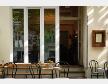 Eingangsbereich eines kleinen Cafés mit hellen Wänden und großen Glastüren, davor eine Terrasse mit runden Holztischen, schwarzen Metallstühlen und kleinen Vasen mit Sonnenblumen. Im Inneren sind Pflanzen und warme Beleuchtung sichtbar.