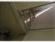 Ein Bereich mit einem Drahtgitterzaun, der an einer Metallkonstruktion befestigt ist, und einer beige gestrichenen Wand im Hintergrund. Elektrische Kabel verlaufen an der Wand, und eine montierte Überwachungskamera ist sichtbar.