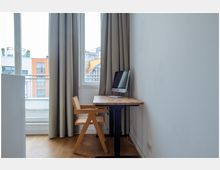 Arbeitsbereich in einer Wohnung mit einem höhenverstellbaren Holztisch, einem Holzstuhl mit geflochtener Sitzfläche und einem Computerbildschirm. Der Raum hat helle Wände, einen Holzboden, bodentiefe Fenster mit beigen Vorhängen und Blick auf umliegende Gebäude.