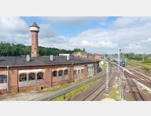 Ehemaliges Bahngelände mit Backsteingebäuden, darunter eine langgestreckte Halle mit kaputten Fenstern, und einem hohen Backsteinturm mit spitzem Dach im Hintergrund. Im rechten Bereich verlaufen mehrere Bahngleise, auf denen auch Züge zu sehen sind, umgeben von einer grünen Landschaft.