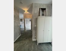 Ein schmaler, moderner Flur mit grauen Wänden und einem grauen Bodenbelag. Rechts steht ein weißer Schrank, und am Ende des Flurs ist durch eine offene Tür ein weiteres Zimmer zu erkennen. Ein Wandlicht beleuchtet den Raum dezent.