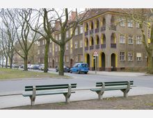 Straßenszene mit Wohnhäusern im Stil des frühen 20. Jahrhunderts, gekennzeichnet durch braune Fassaden mit gelben und violetten architektonischen Elementen sowie Arkaden. Im Vordergrund befinden sich eine Straße mit parkenden Autos, mehrere Bäume und zwei grüne Parkbänke.