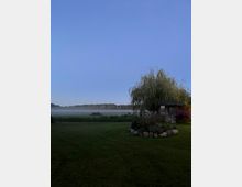 Weitläufige Rasenfläche am Rand einer nebligen Wiese oder eines Feuchtgebiets, mit Baumreihe am Horizont unter klarem Himmel. Rechts steht ein kleines Holz-Gartenhäuschen neben einer großen Trauerweide, umgeben von einem Blumenbeet mit Steinen.