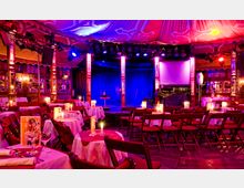 Ein festlich beleuchteter Veranstaltungssaal mit Bühne, umgeben von reich verzierten Holzsäulen und einem drapierten Stoffdach. Im Vordergrund sind runde Tische mit weißen Tischdecken, brennenden Kerzen und Stühlen angeordnet.