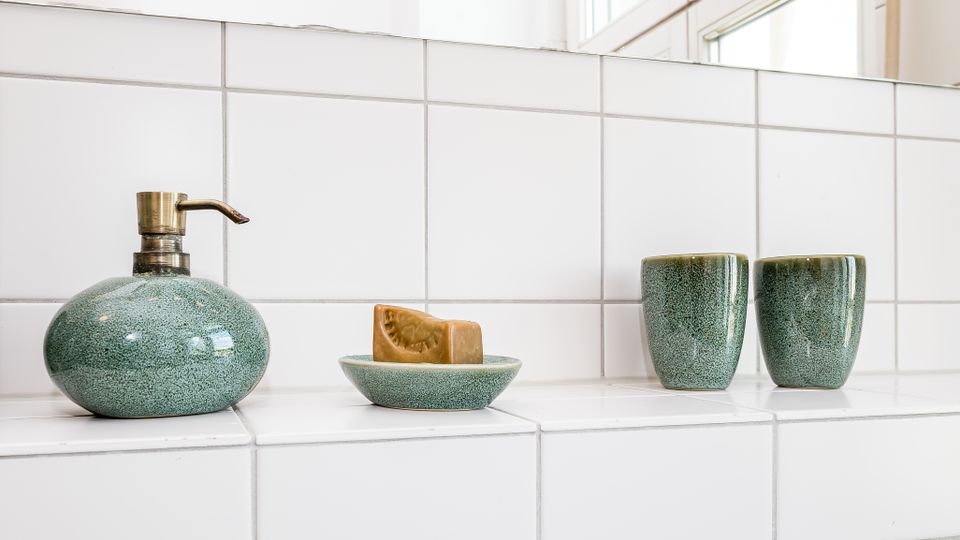Weiße, geflieste Badezimmerablage unter einem Fenster mit grüner Keramik-Seifenspenderflasche, Seifenschale mit Stückseife und zwei passenden Bechern. Die rechteckigen Wand- und Ablagefliesen prägen das helle, minimalistische Ambiente.