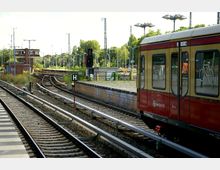 Eine S-Bahn-Station mit mehreren Gleisen und einem einfahrenden Zug. Im Hintergrund sind grüne Bäume, ein kleines Backsteingebäude und Signalleuchten sichtbar.