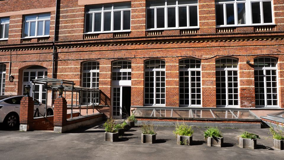 Innenhof vor einem mehrstöckigen Backsteingebäude mit großen, weiß gerahmten Fenstern und Rundbögen im Erdgeschoss. Im Vordergrund stehen Betonpflanzkübel, daneben befindet sich ein gläsernes Vordach und ein langes Glasdach über einem Lichtschacht.